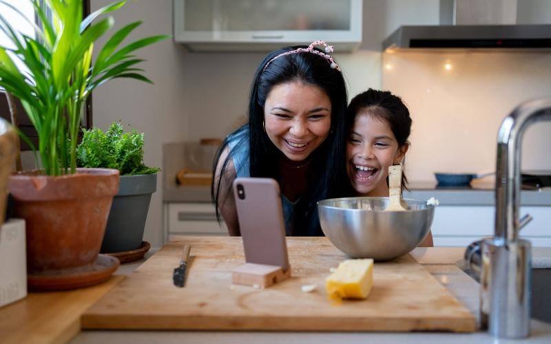 mother and young daughter smile at their phone as they cook in the kitchen
