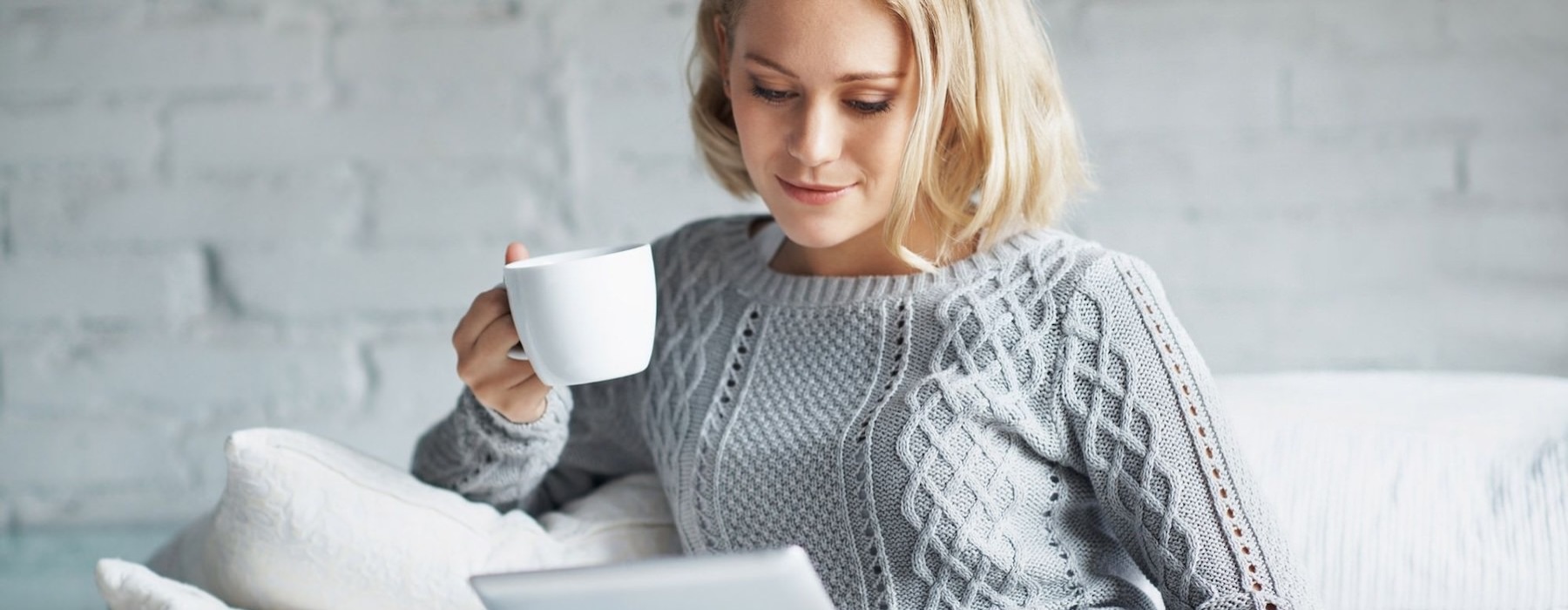 a woman sits on a couch with a cup of coffee and looks at her tablet